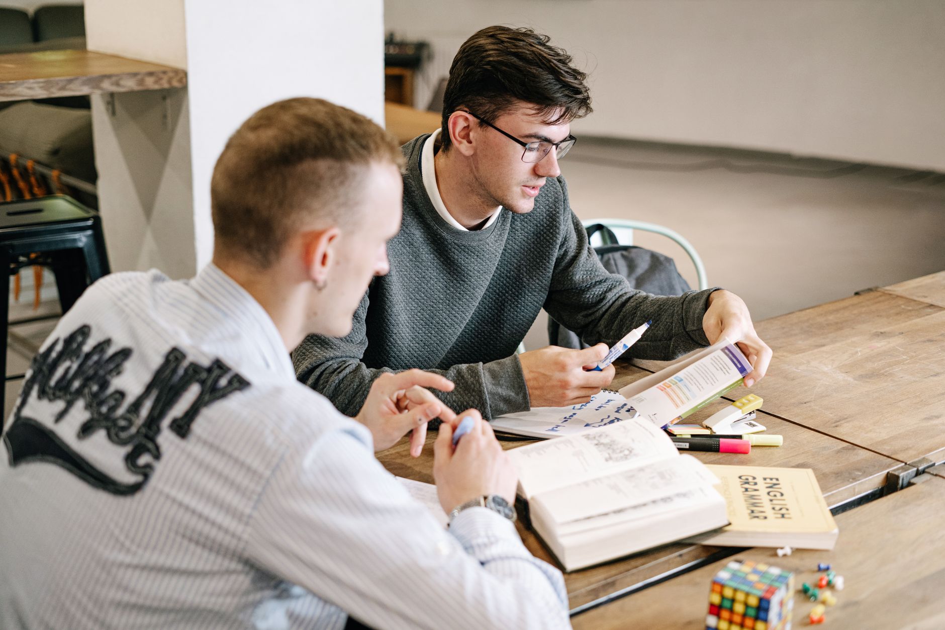 English for University Studies — Two young men studying together, focusing on English grammar books in a modern indoor setting.