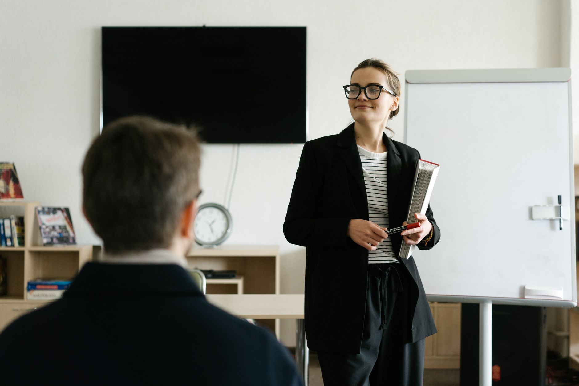 Personal Effectiveness — Caucasian female teacher in black blazer holding books, presenting to a student in a classroom.