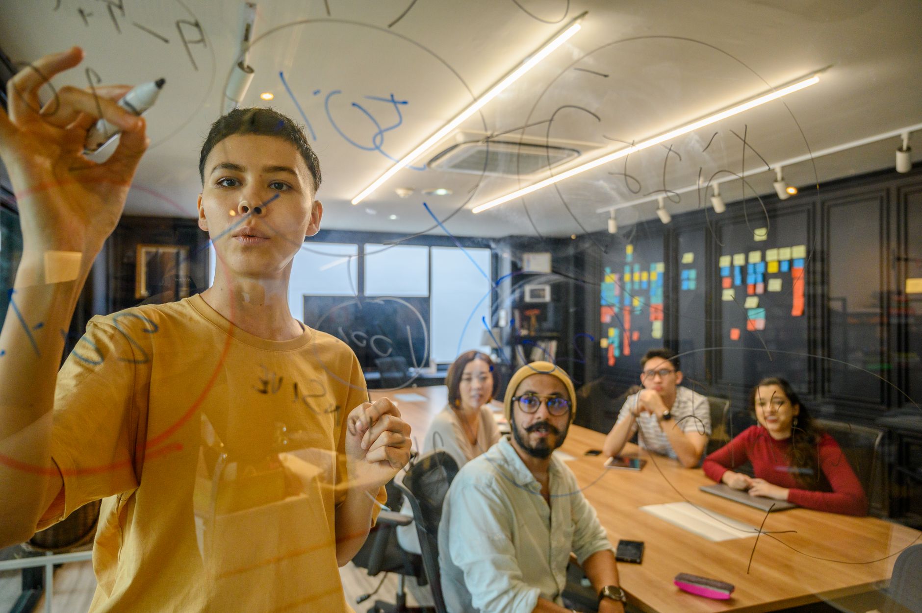 Innovation in Project Management — A team of young professionals working together in a modern office setting, brainstorming and writing on a glass board.