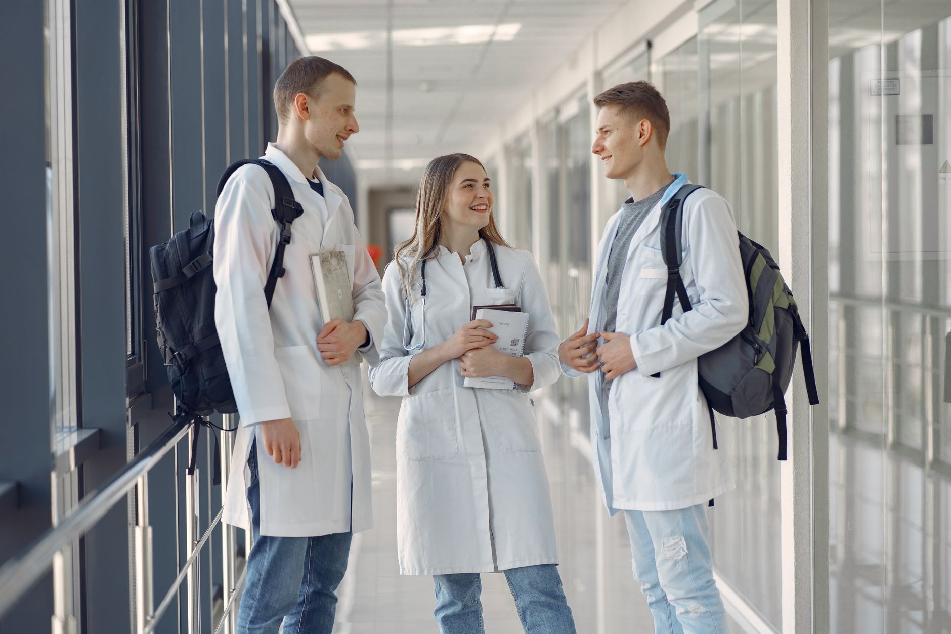 Implement Person-Centred Approaches — Three young medical professionals in lab coats talking in a hospital hallway.