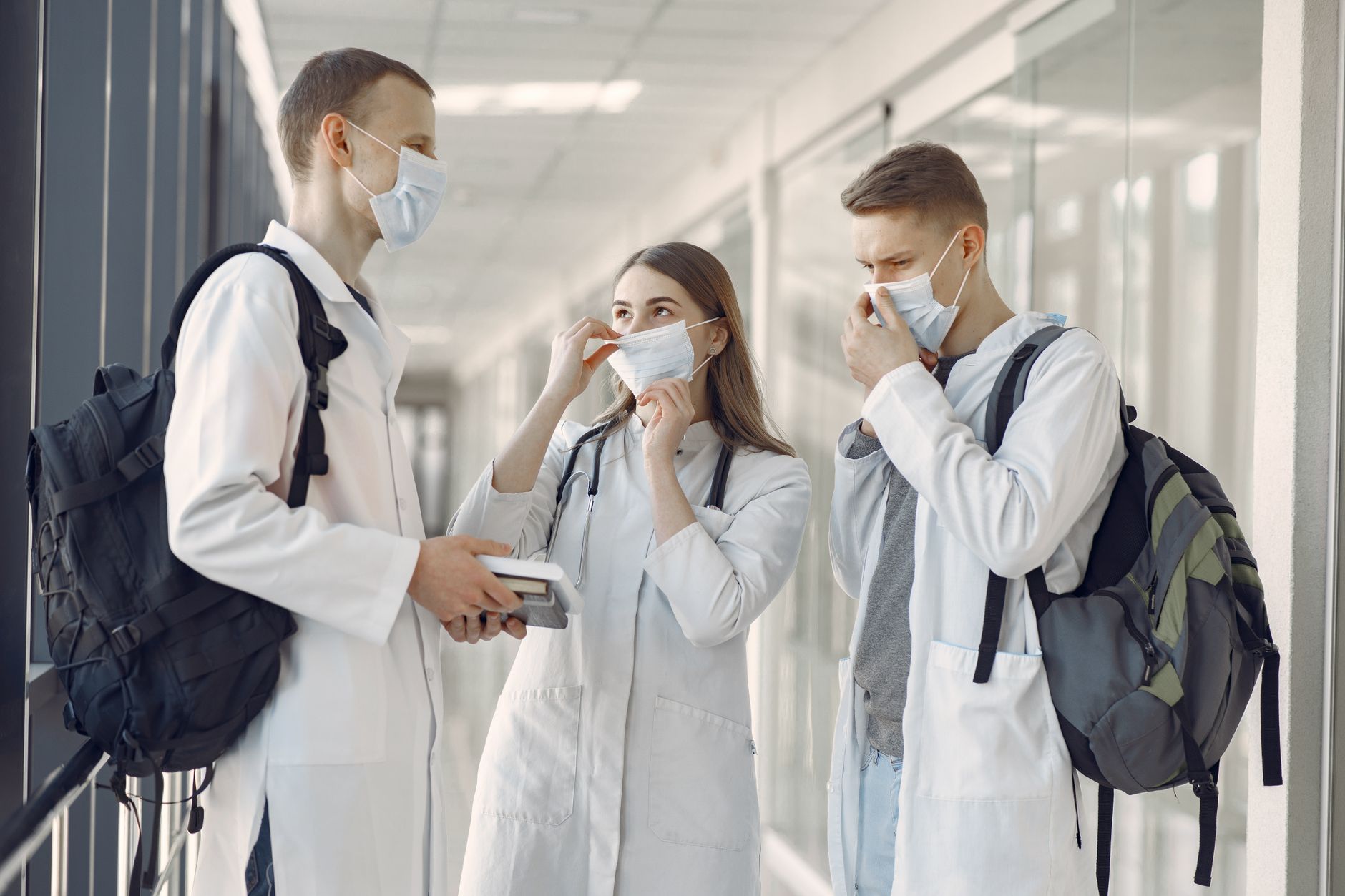Safeguarding and Protection in Care Settings  — Three young medical professionals discuss in a hospital corridor wearing protective masks.
