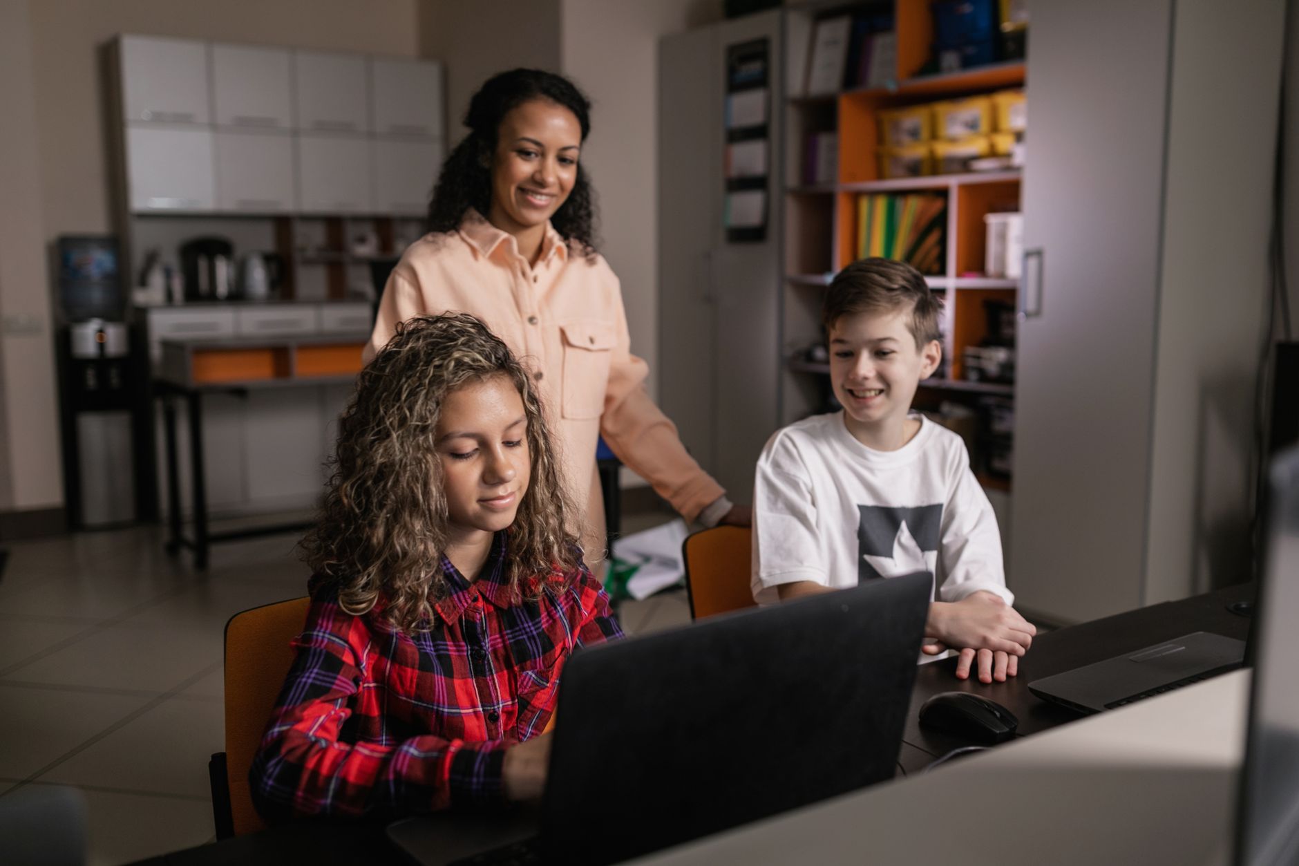 Contribute to Support of Positive Risk- taking for Individuals — A teacher supervising two smiling teenagers working on laptops in a classroom setting, focusing on technology and learning.