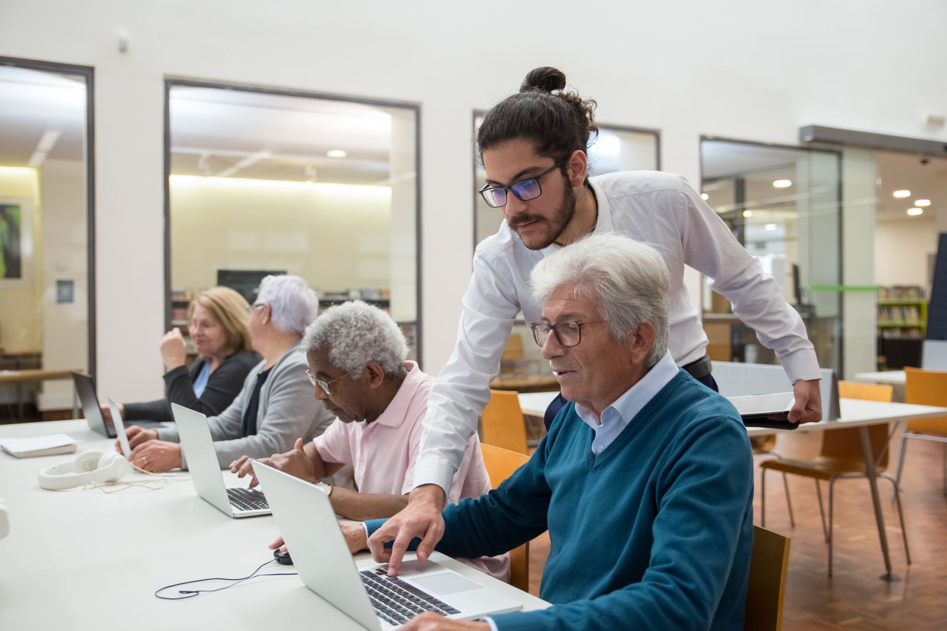 Understand How to Support Individuals to be Part of a Community — A teacher guiding senior adults in a library, using laptops for learning and support.