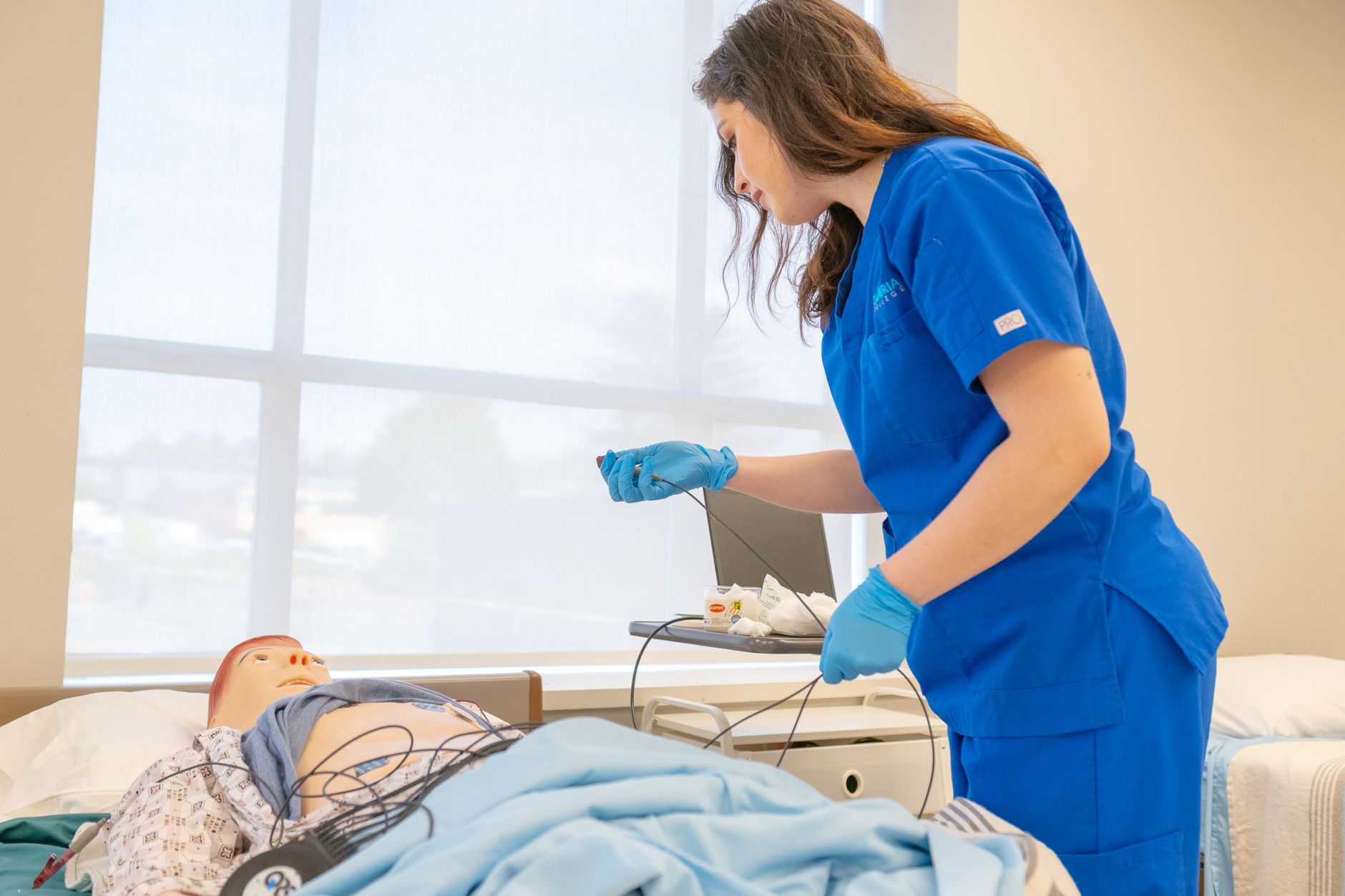 Understanding Stroke Care — A nursing student in blue scrubs attends to a medical mannequin in a training room.
