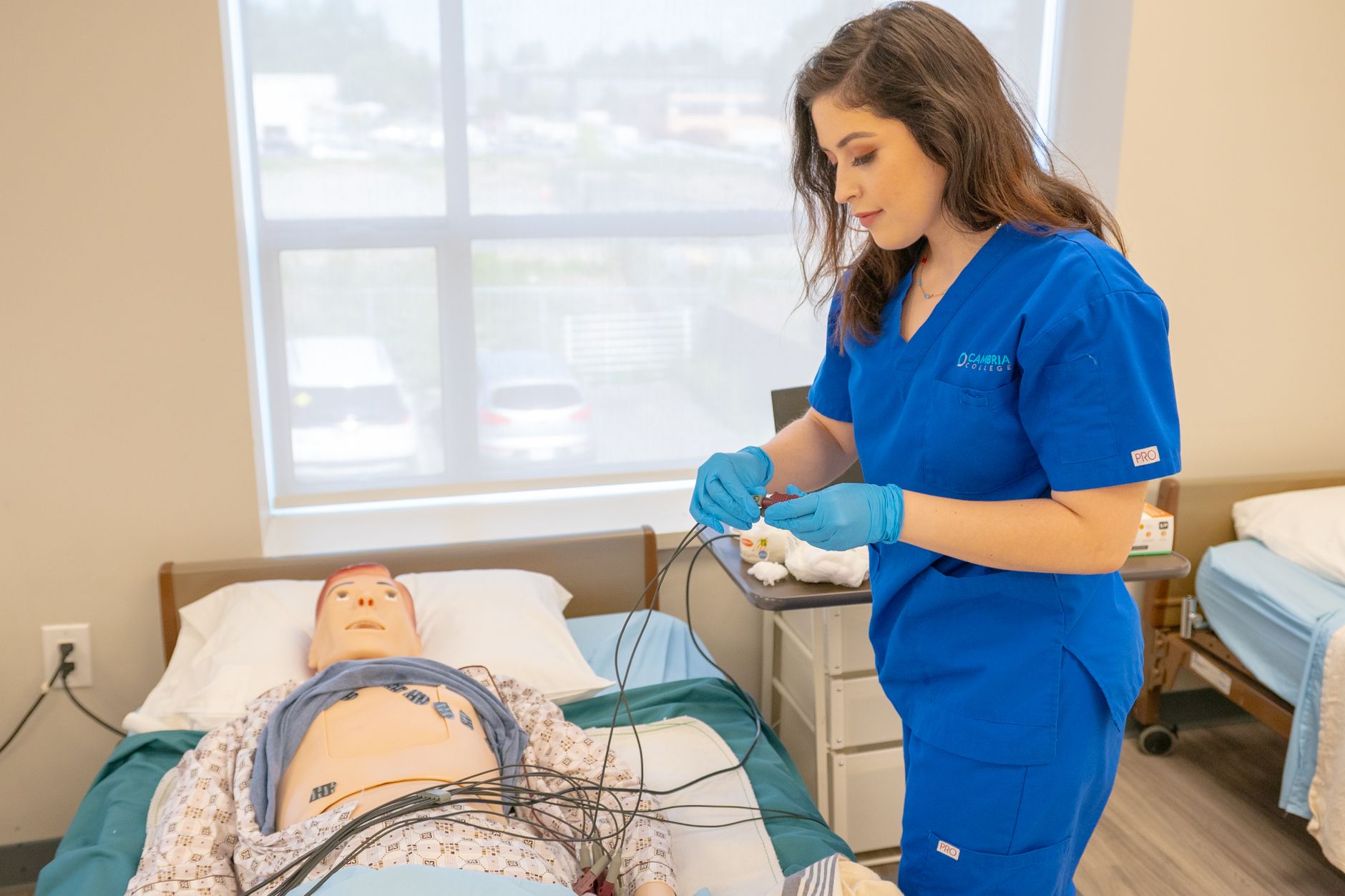 Understanding Dementia Care — Medical student in blue scrubs practicing procedures on a patient training manikin in a clinical setting.