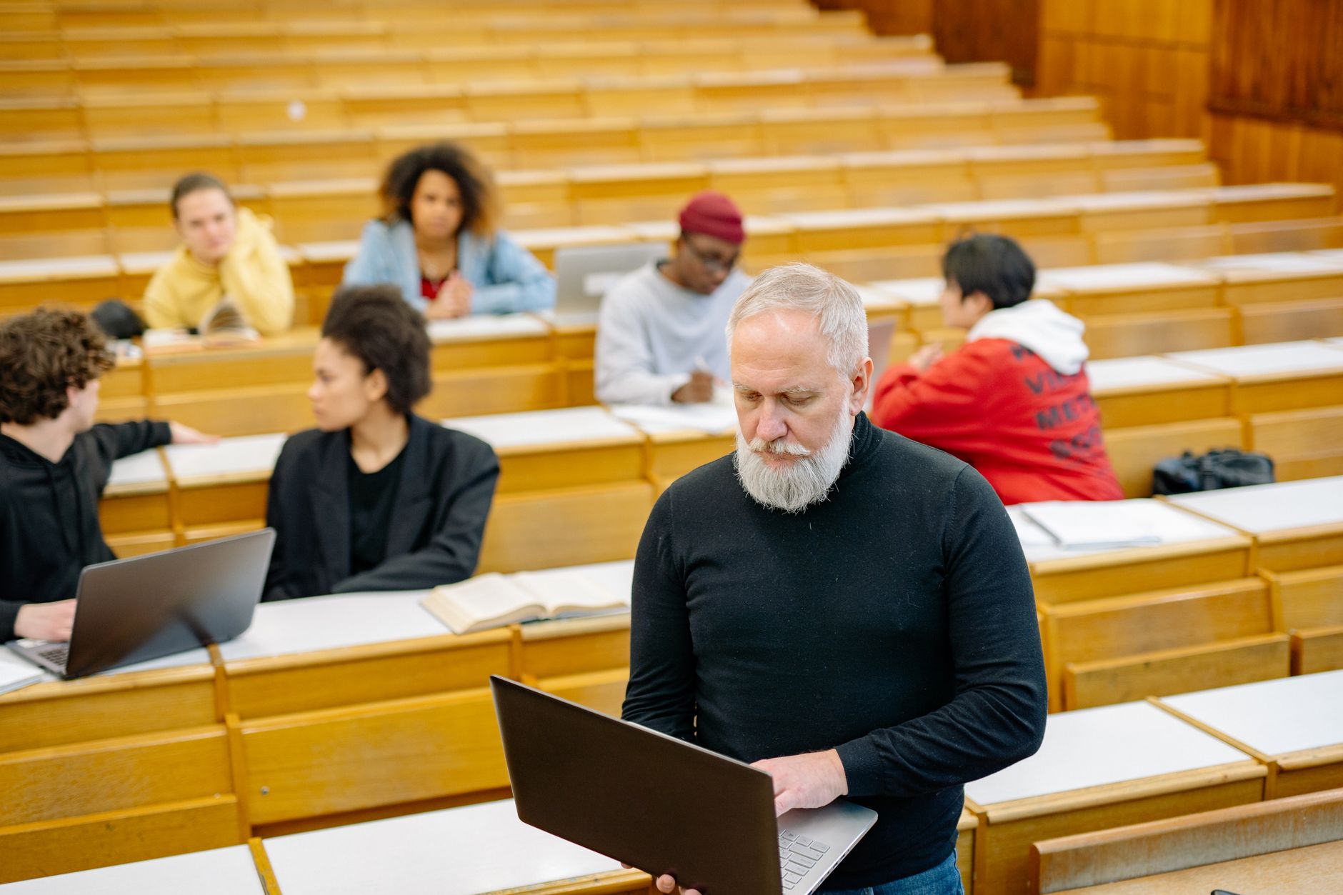 The Diversity of Addiction — Mature teacher interacting with diverse students using laptops in a spacious lecture hall setting.