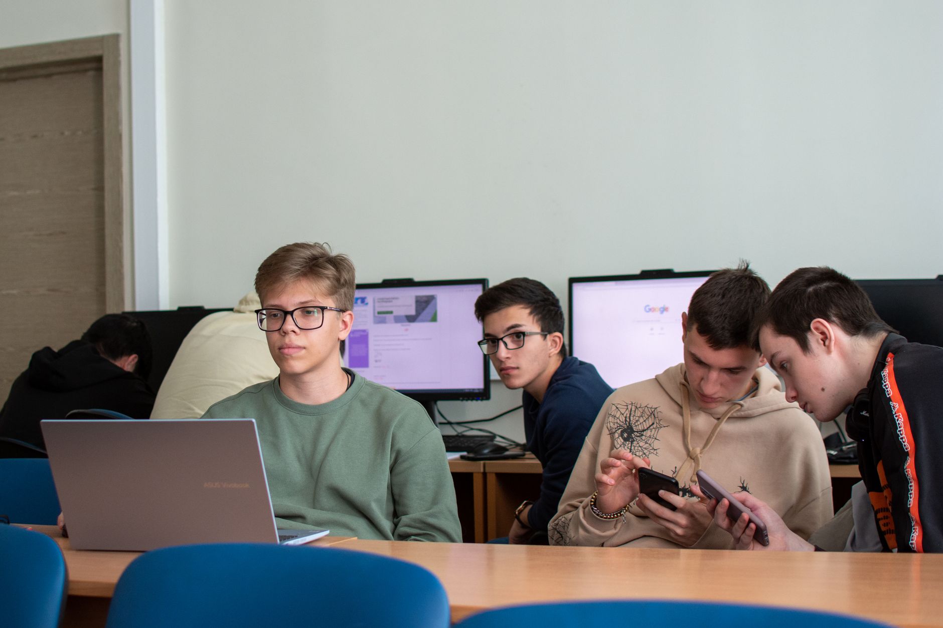 Information Technology and IT Ethics — Five teenagers engaged with laptops and smartphones in a modern classroom.