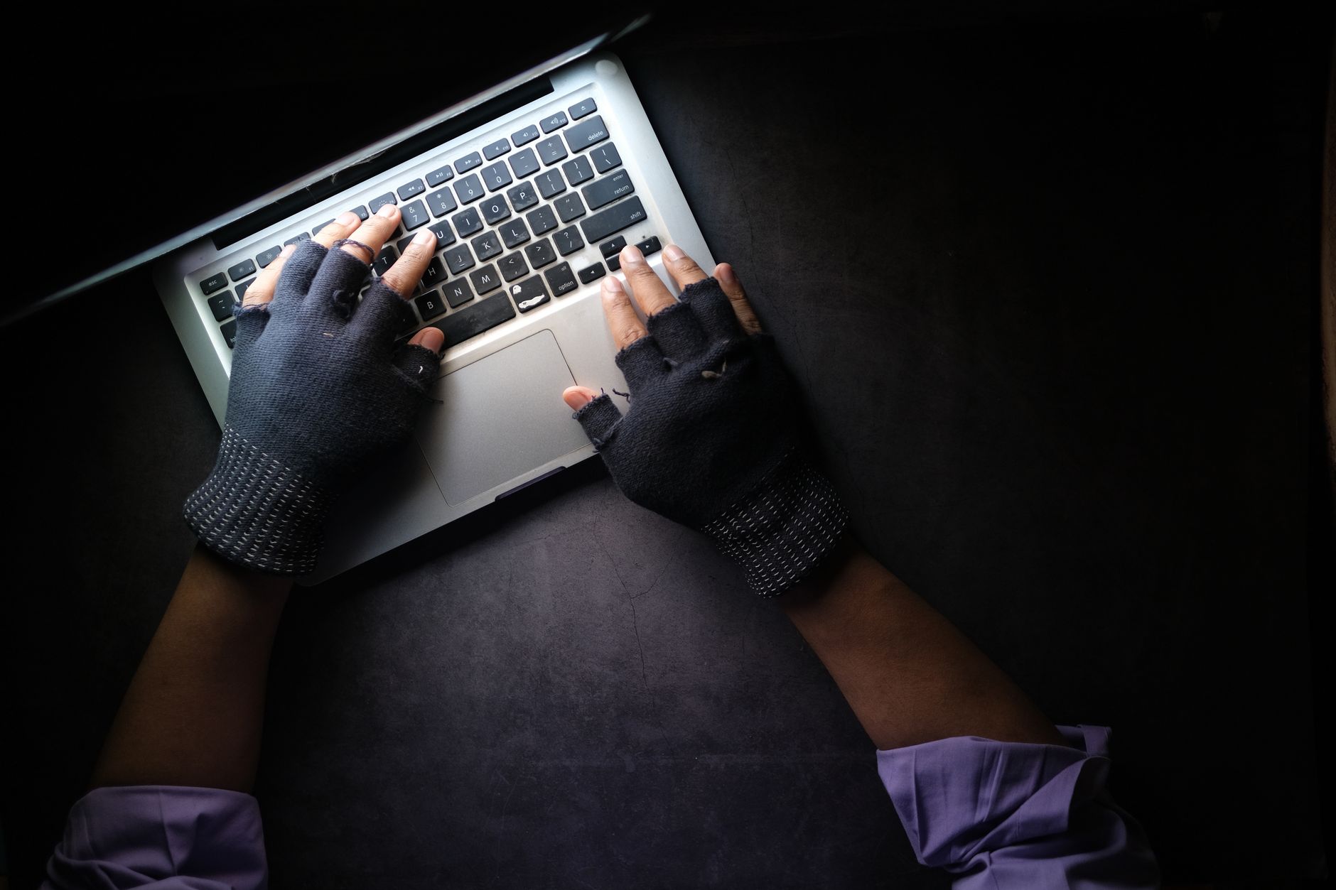Cyber Security Threats and Risk: Banking and Finance — Hacker in fingerless gloves typing on laptop keyboard from above in a dark setting.