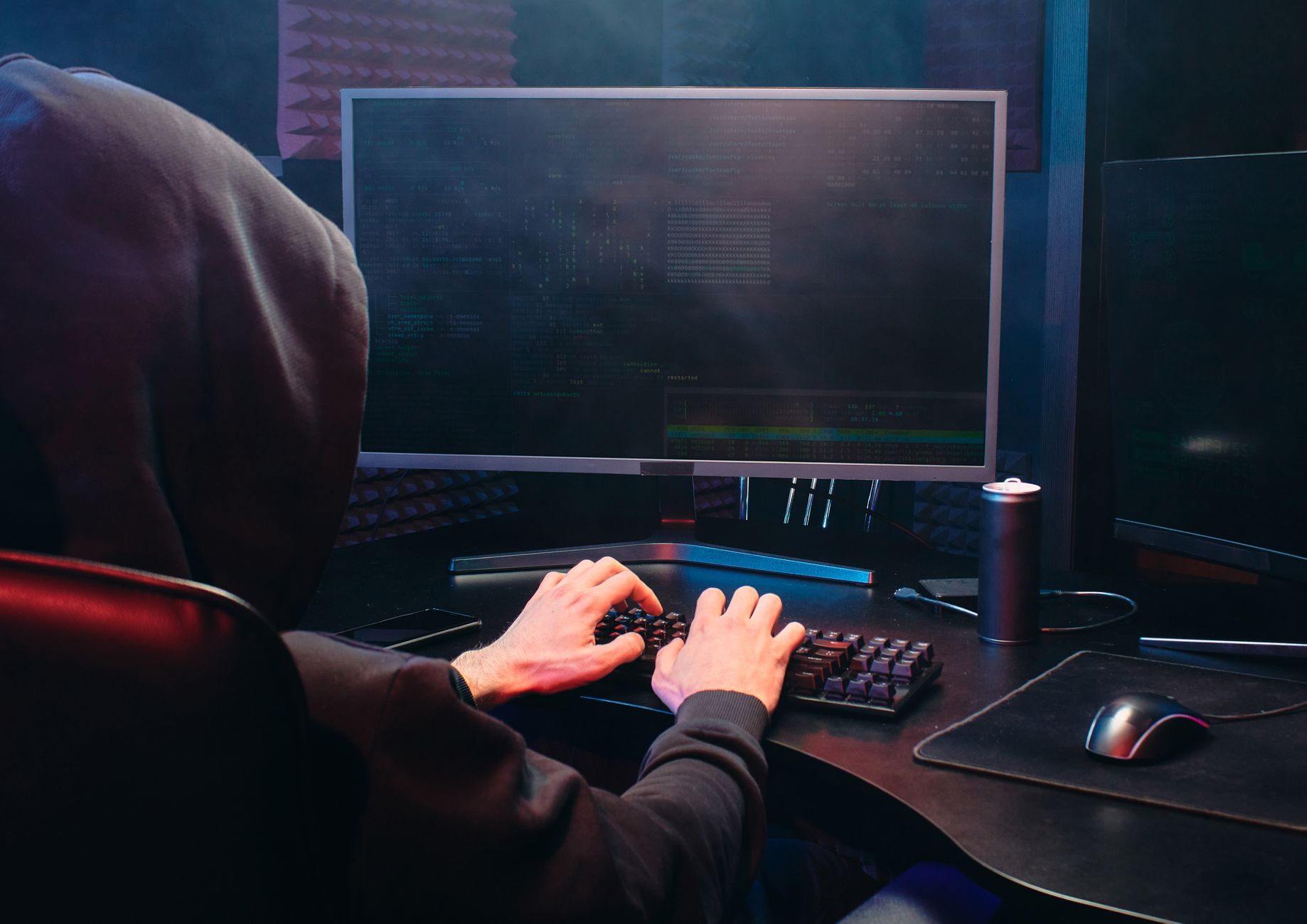 Cyber Security Threats and Risk: Banking and Finance — A person in a hoodie sits at a computer screen, engaged in coding or hacking activities.
