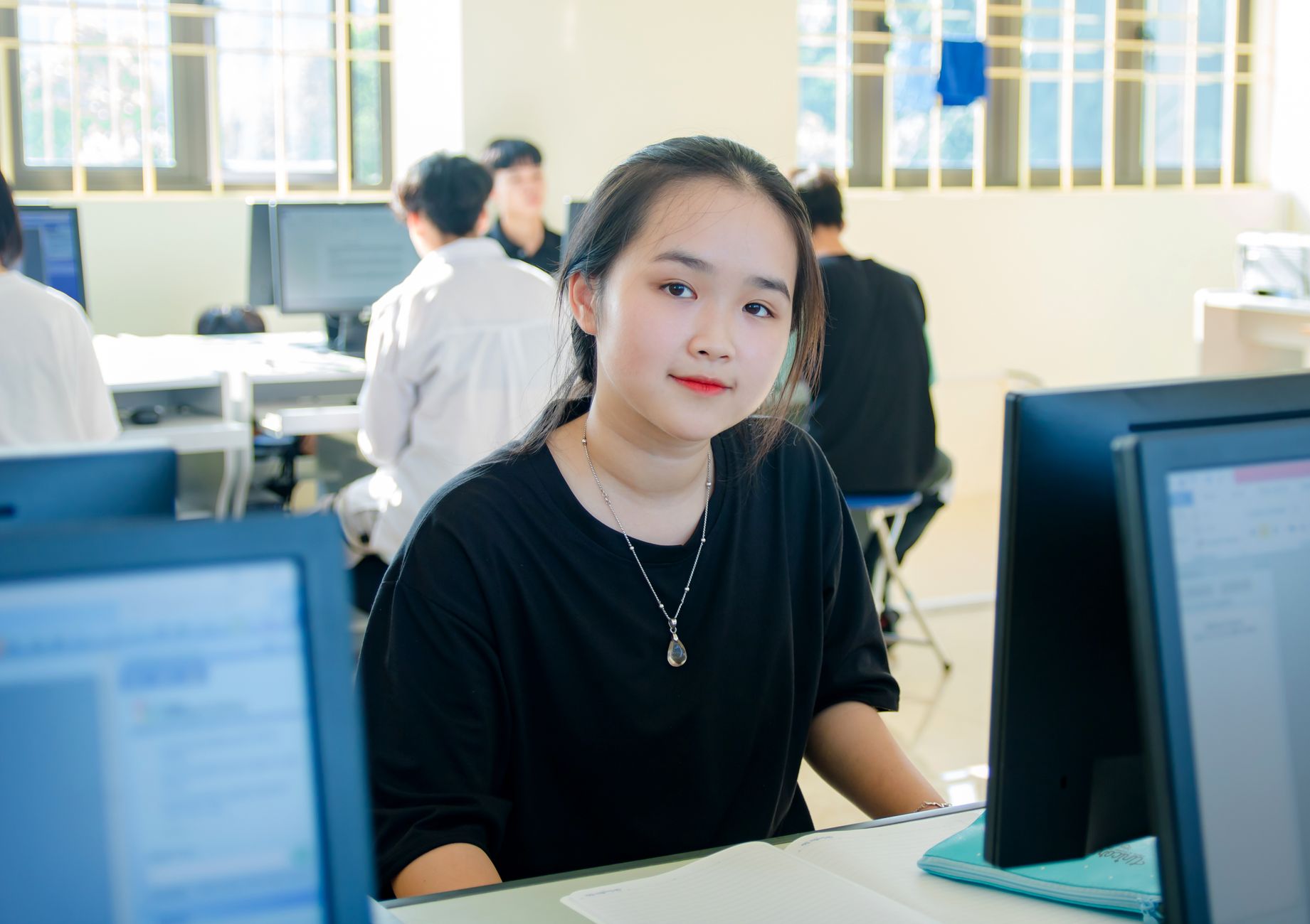 Working relationships with children, young people and adults. Schools as business organisations.Support for learning activities — A young woman sits at a desk in a computer lab, smiling into the camera.