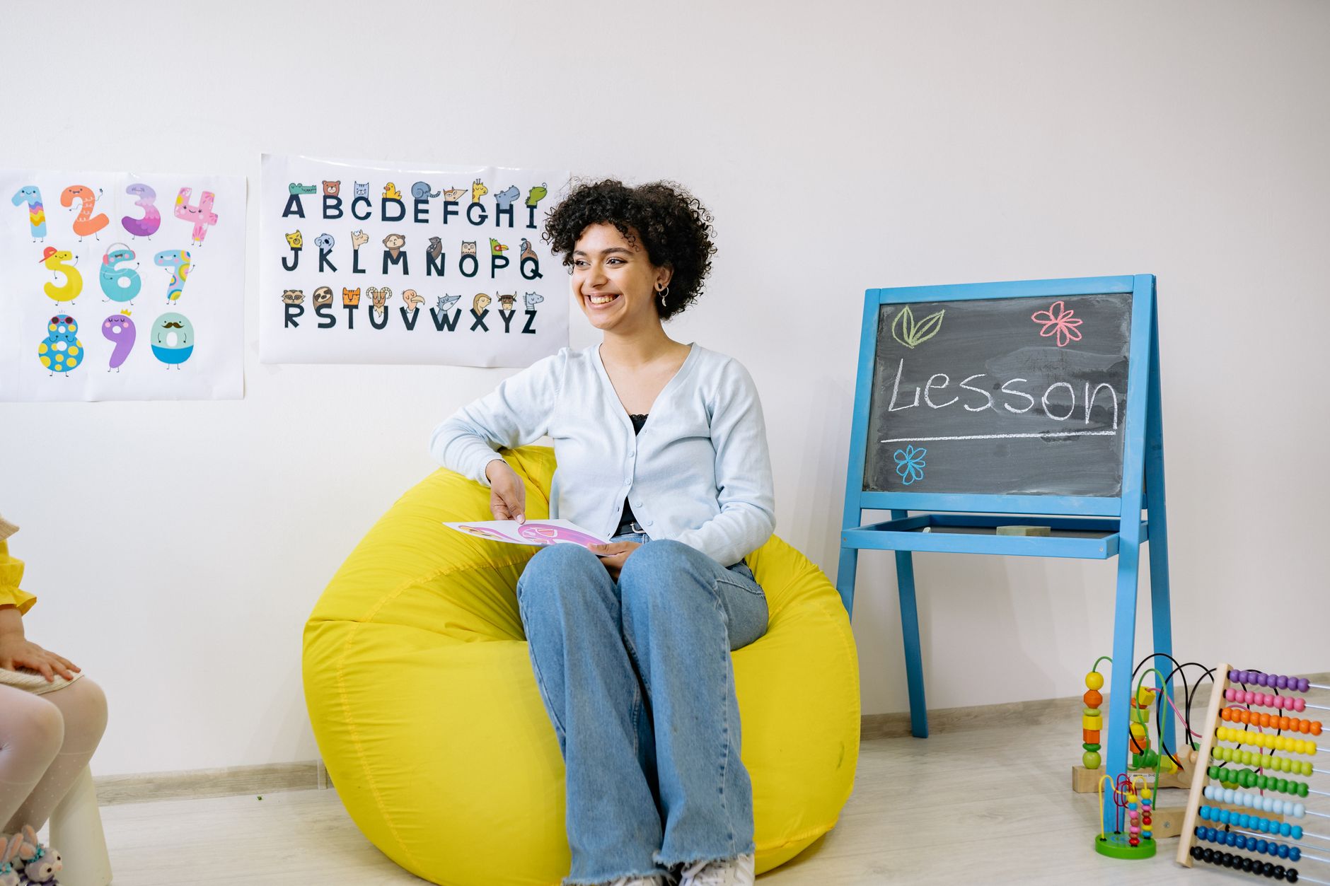 Teaching Materials and Aids and Classroom Management Strategies — Smiling teacher sitting in a classroom, holding a book and teaching preschool students.