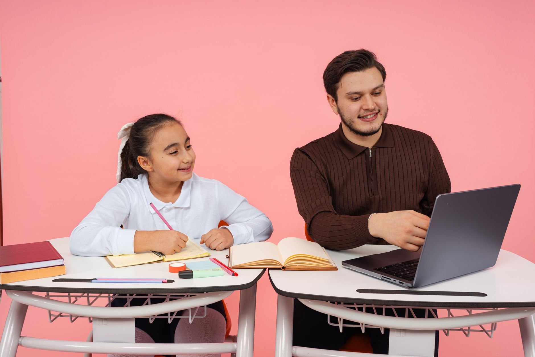 Designing Learning Materials — A teacher guides a student in a classroom with books and a laptop on a pink background.