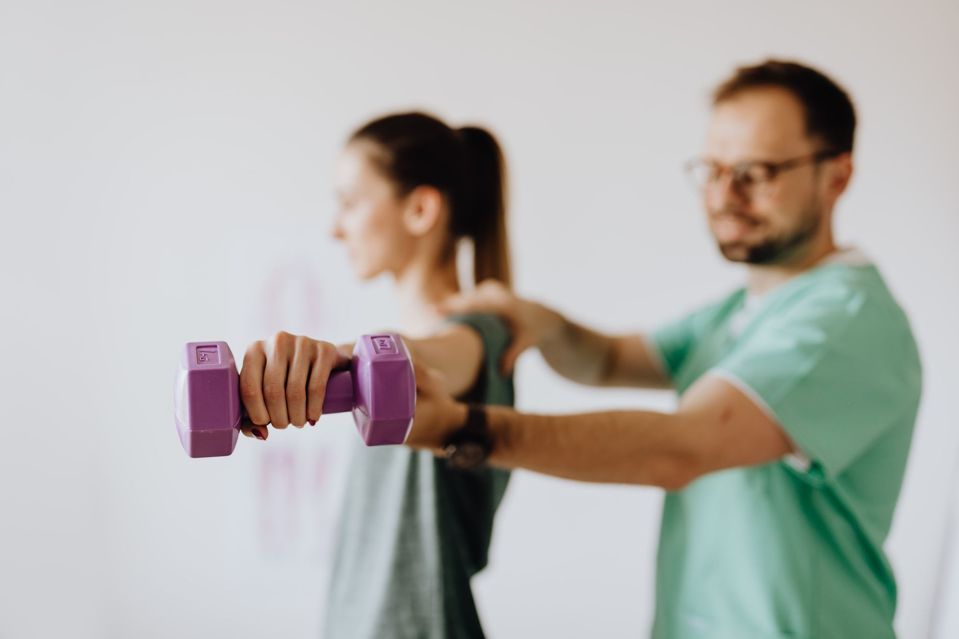 The Foundations of Health and Wellness — Side view of professional orthopedist in uniform and eyewear helping fit woman reaching arm with dumbbell in doctor office on blurred background