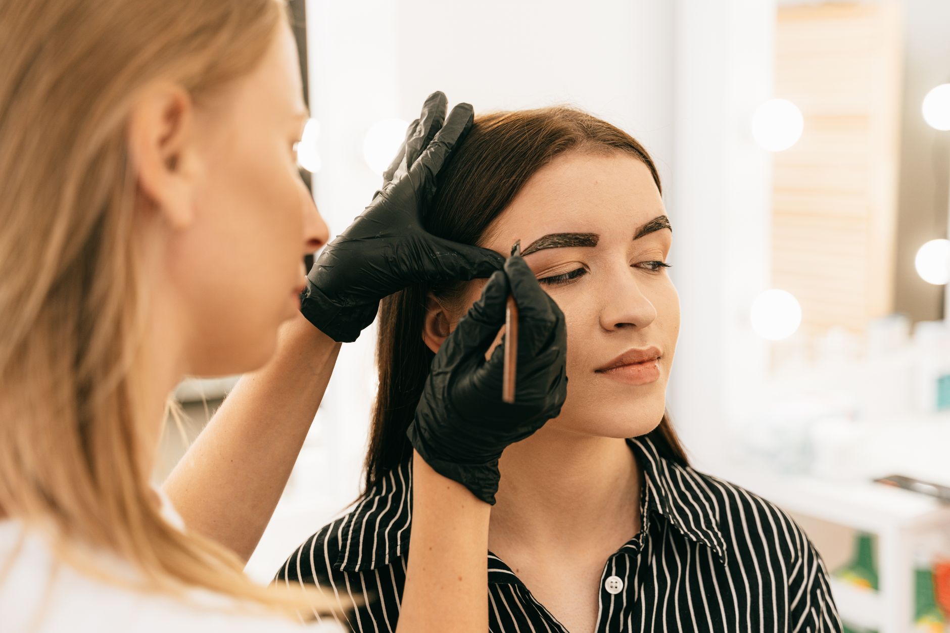 units – 35 credits: — A professional makeup artist shapes a woman's eyebrows in a bright beauty studio.