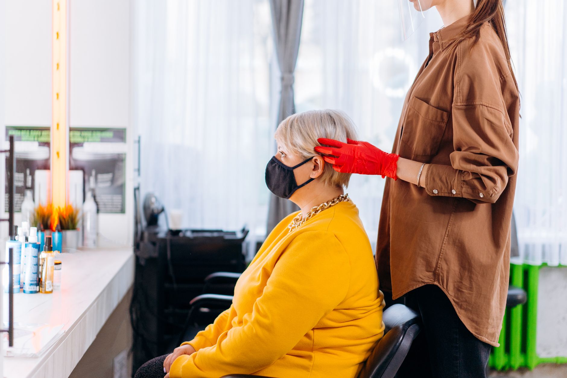 Health, safety and hygiene for aesthetic procedures — Side view of female client and hairstylist in masks and casual clothes with gloves near table with professional equipment and mirror in bright salon