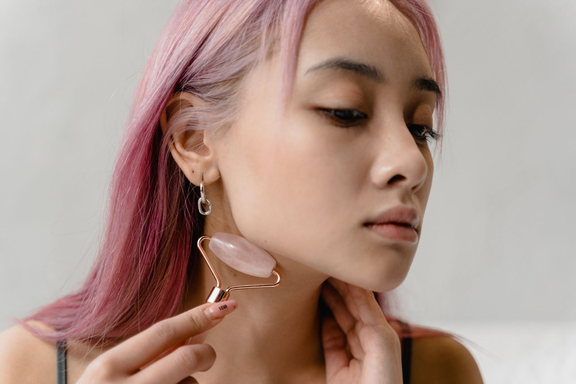 Temporary and Reversible Dermal Fillers to the Face and Neck — A young woman with pink hair uses a facial roller for a soothing skincare ritual.