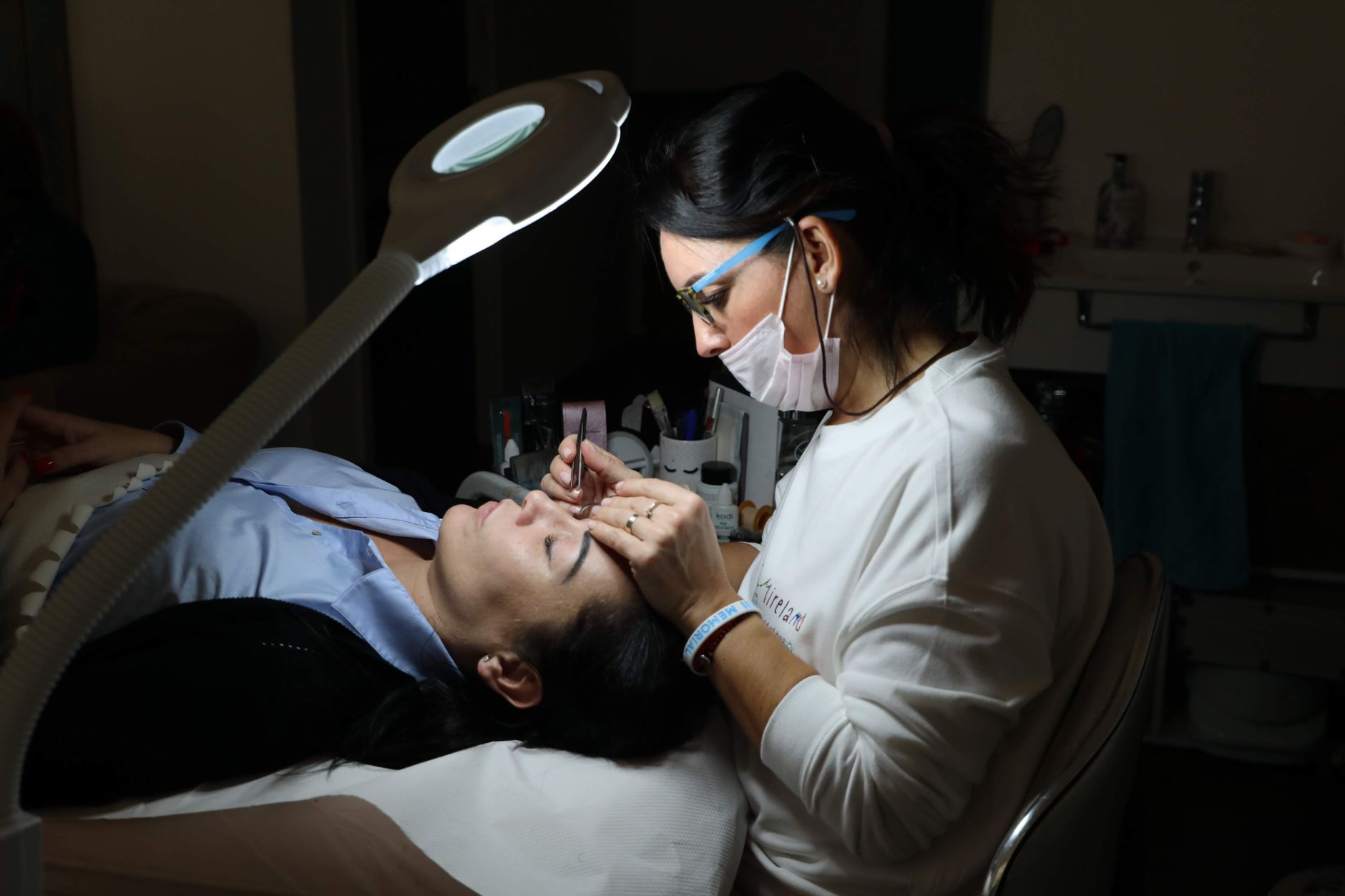 Client consultation — A beautician provides professional eyebrow care to a client in a salon, under focused lighting.