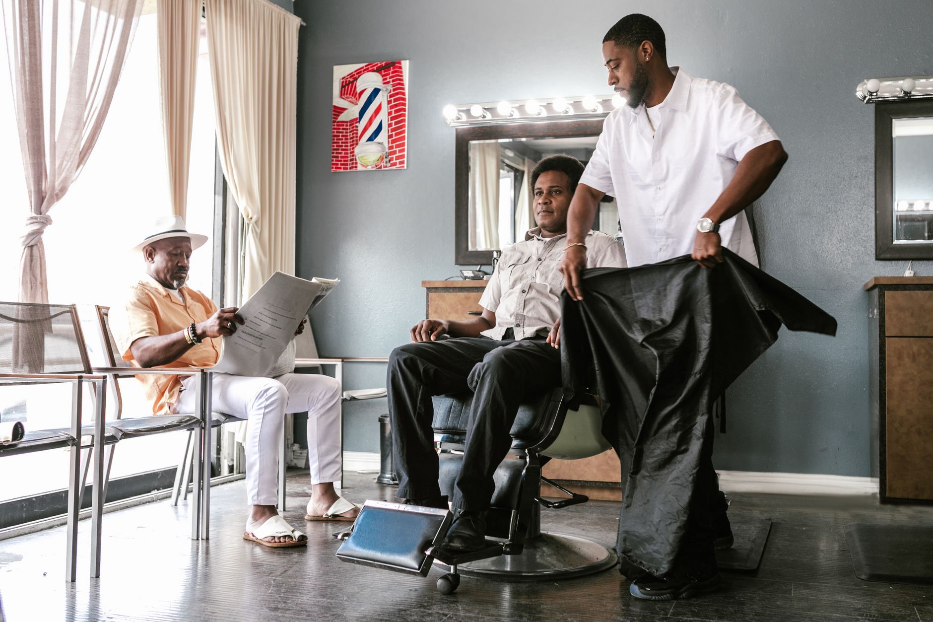 Consultation for barbering services — A man receives a haircut in a barber shop while another customer reads a newspaper.