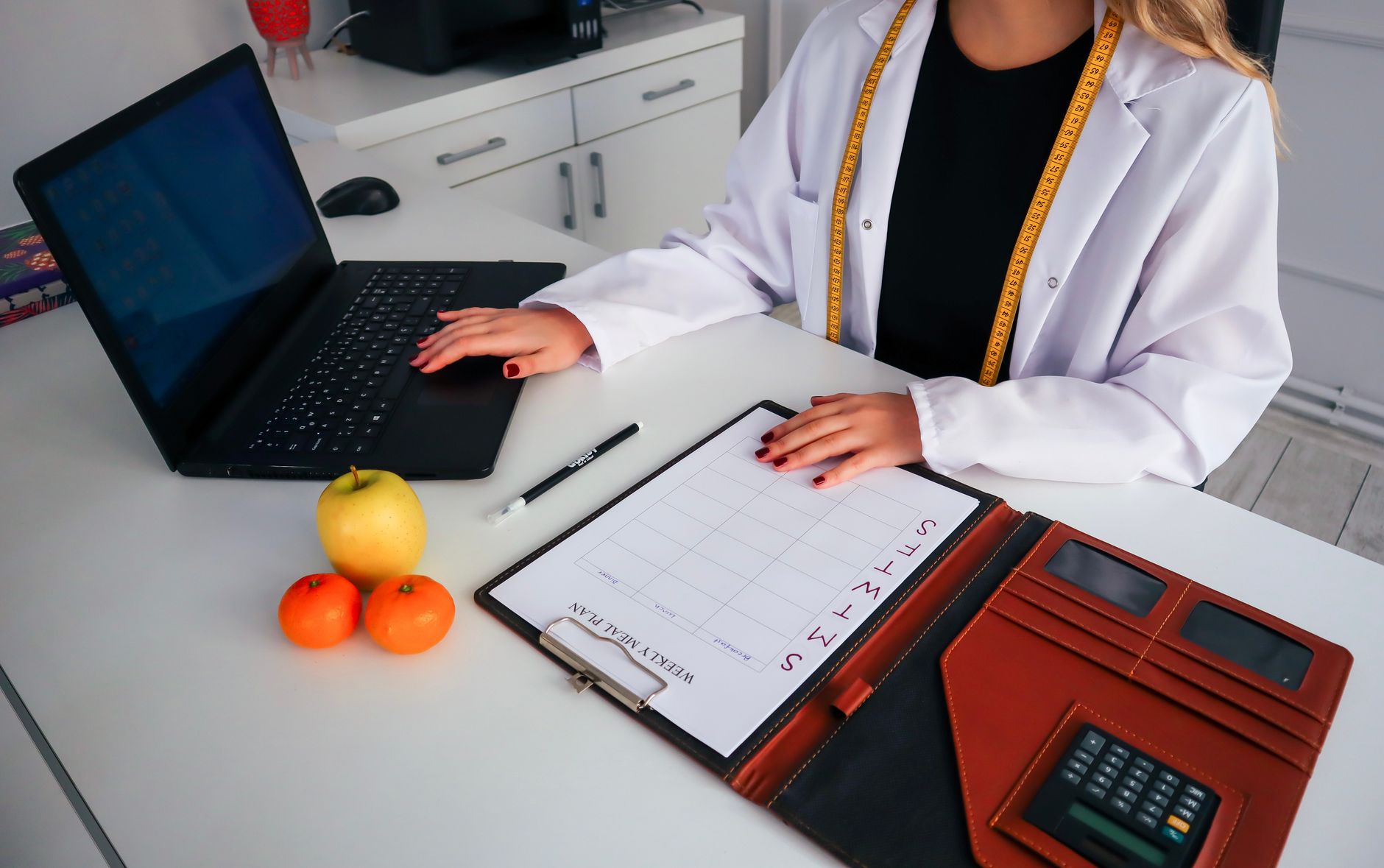 Health and Nutrition – Nutrition and Health Coach Practice — Dietitian working on meal plan with laptop, fruits, and calendar for health consultation.
