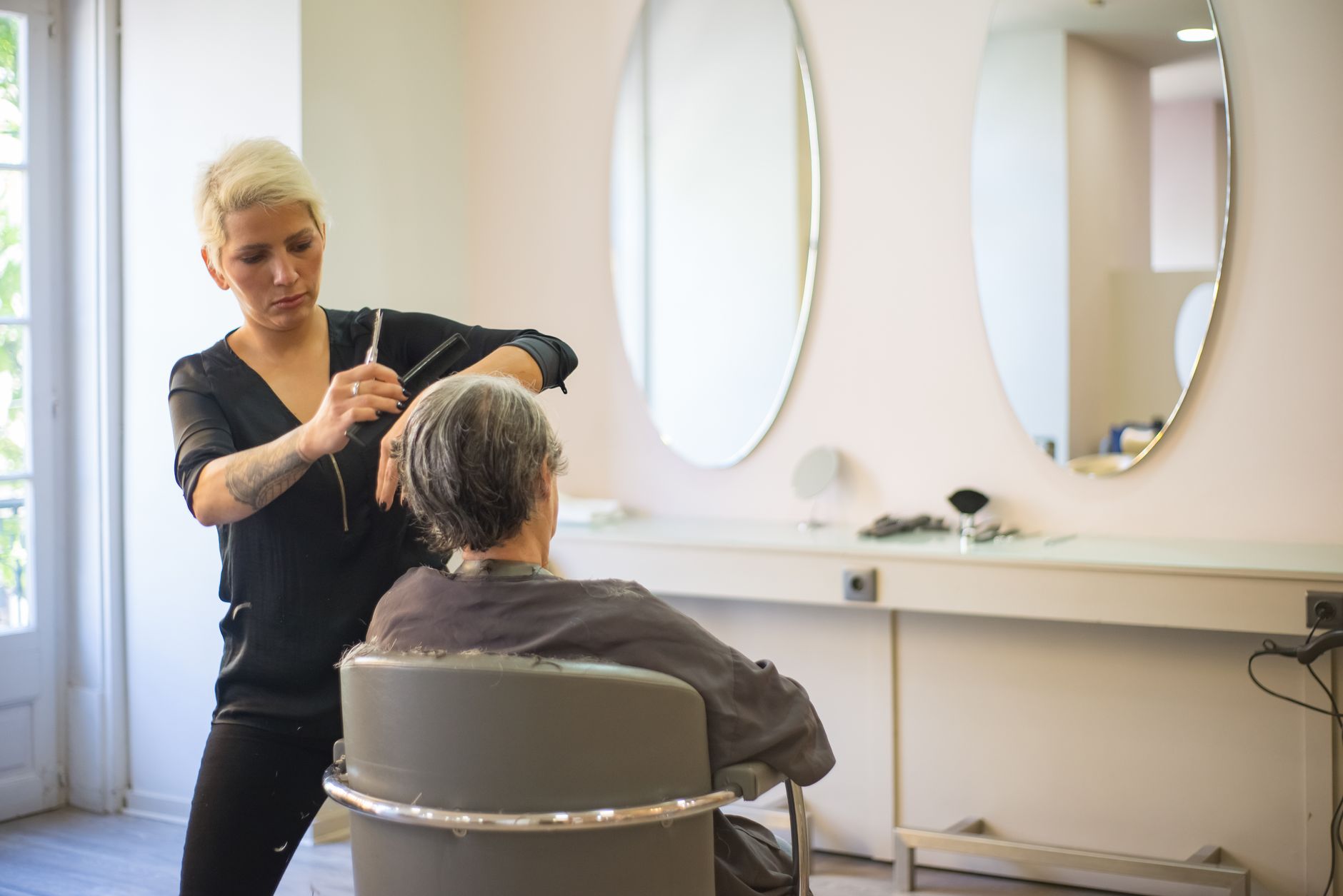 Freelance worker — A hairdresser skillfully cuts an elderly woman's hair in a modern salon with mirrors and a comfortable barber chair.