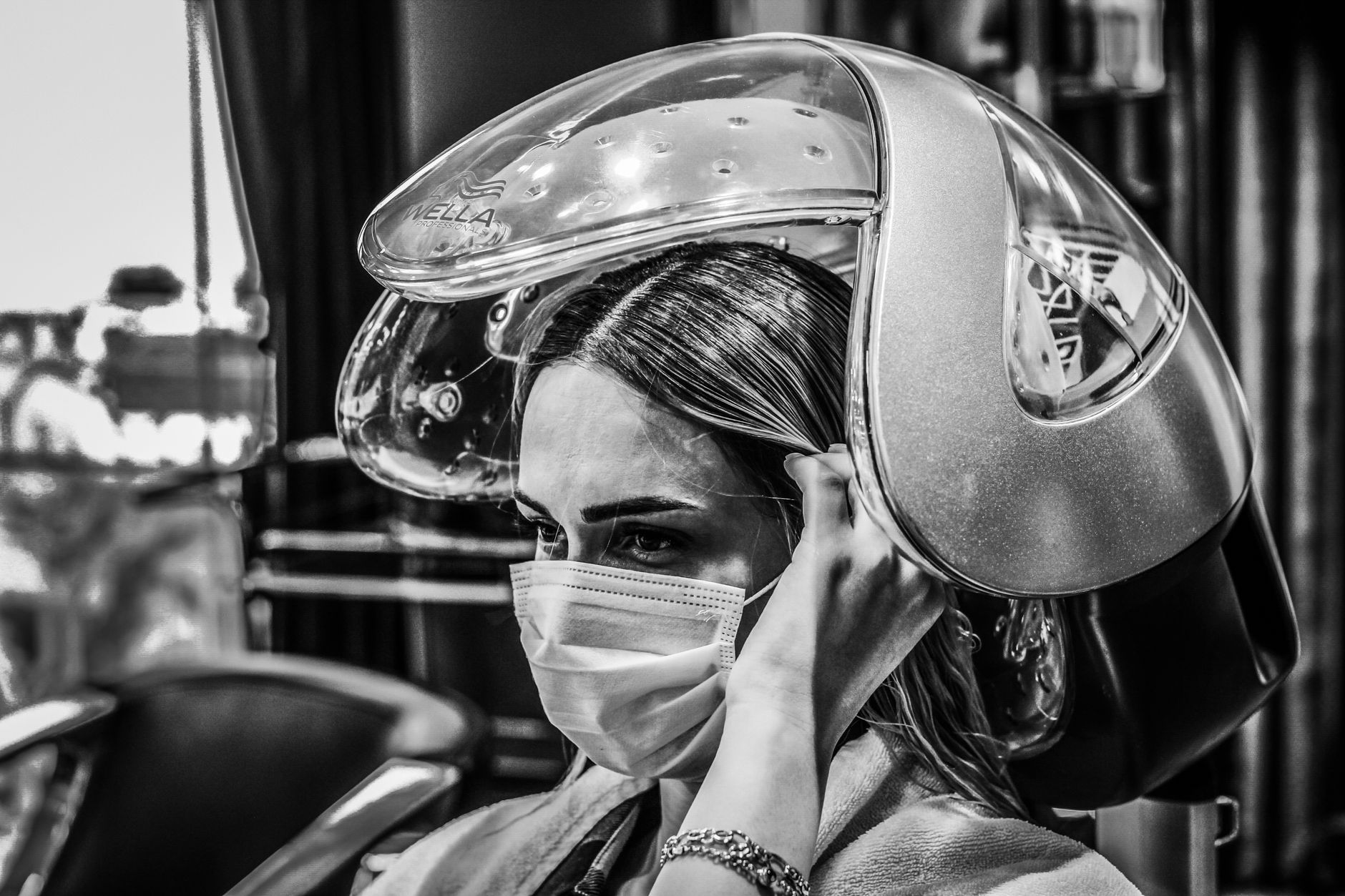 Implement health, safety, and hygiene — A woman in a hair salon wears a face mask under a hair steamer for safety.
