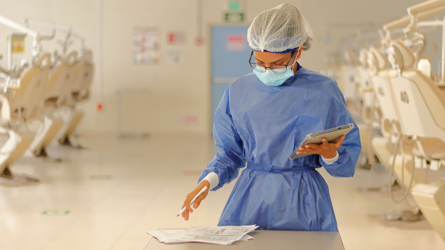 Implement health, safety, and hygiene — A healthcare professional in blue protective gear reviews documents in a medical clinic.