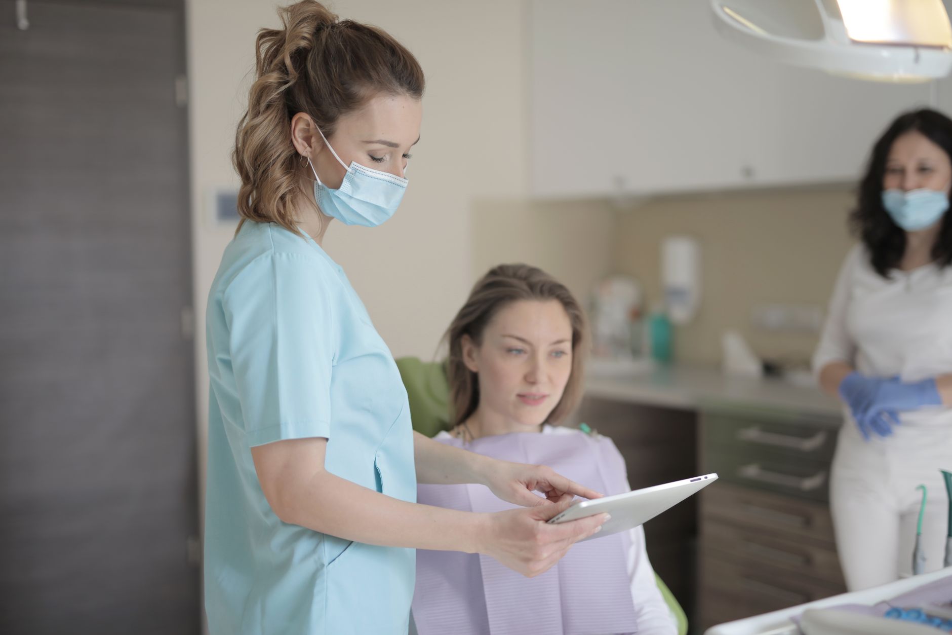Implement health, safety, and hygiene — Young female dentist in mask and uniform using tablet while talking with patient before treatment in modern dental clinic in hospital