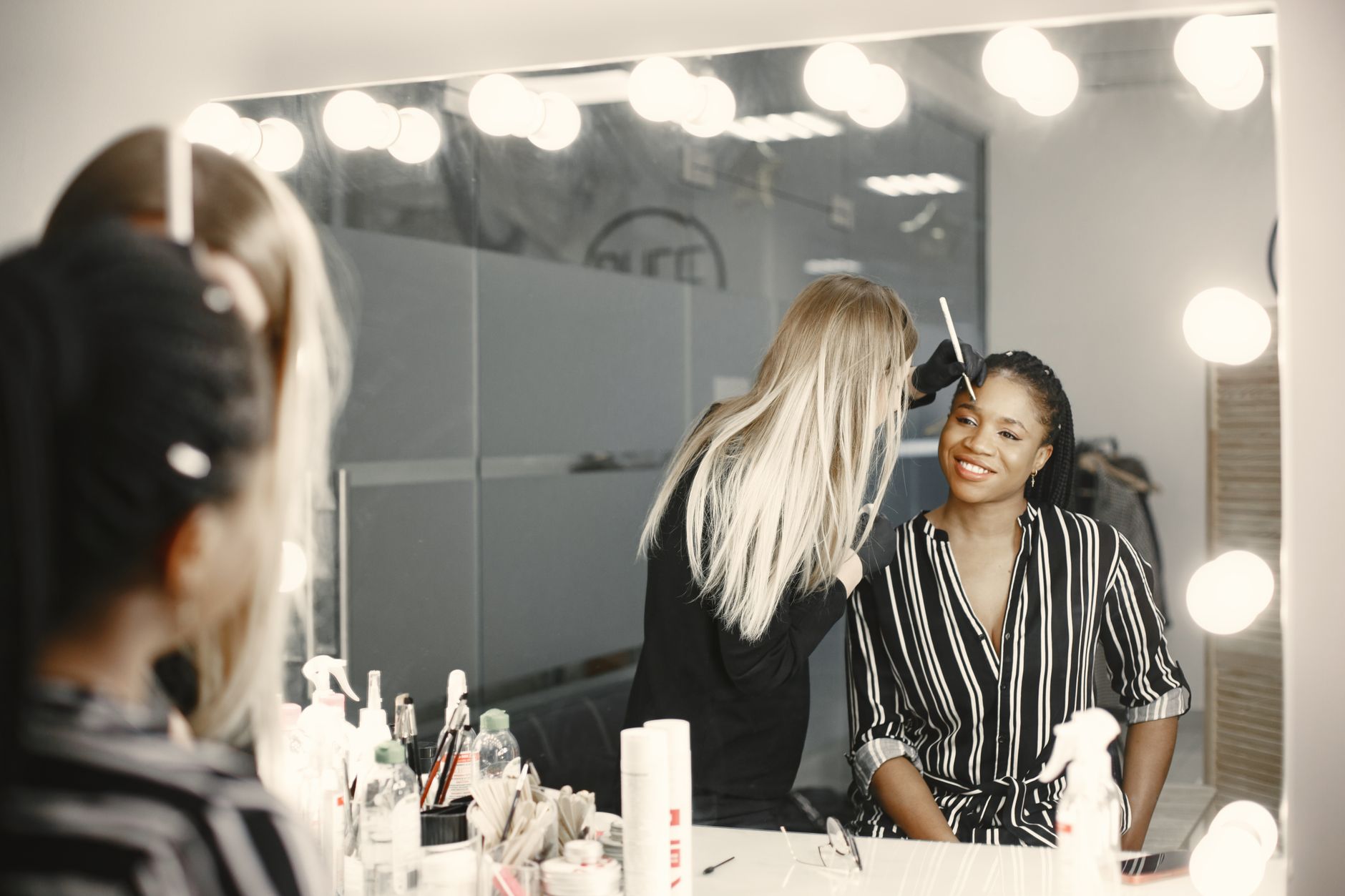 Creatively set, dress, style, and finish hair — A professional makeup artist applies makeup to a smiling woman in a well-lit backstage setting.