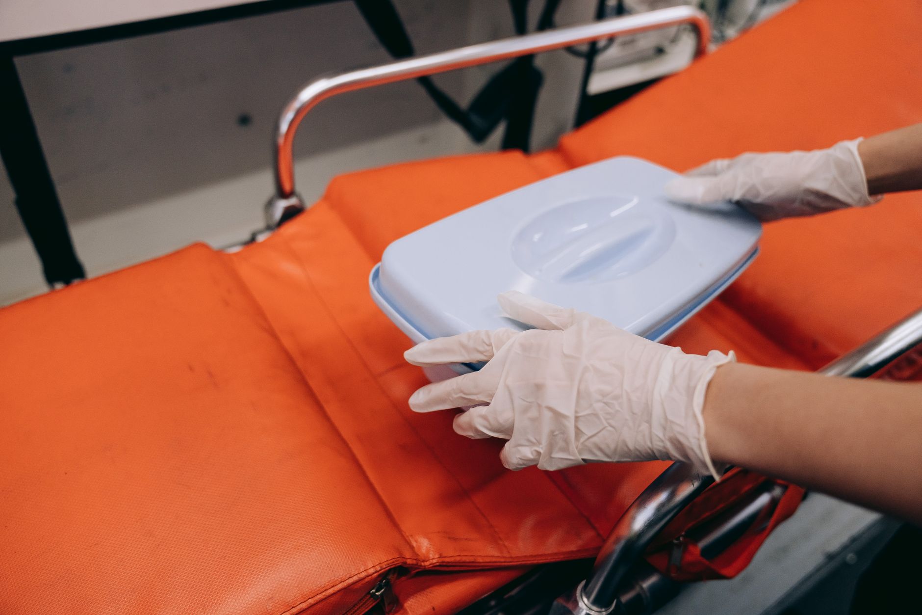 Implement health, safety, and hygiene — Focused shot of gloved hands handling a first aid kit on an orange stretcher in a medical setting.