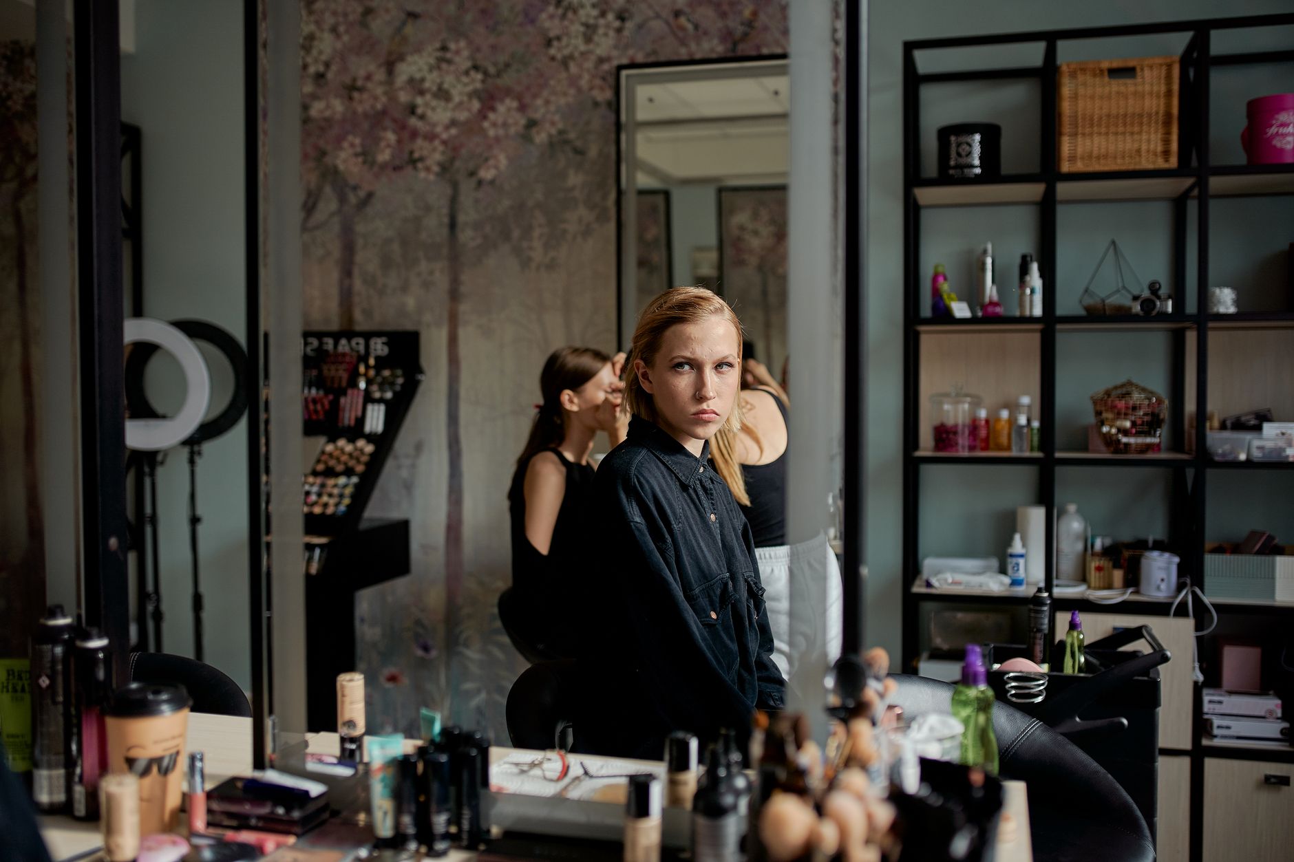 Implement health, safety, and hygiene — Side view of young attentive blond lady in black apparel looking in mirror near table with assorted cosmetic products in salon